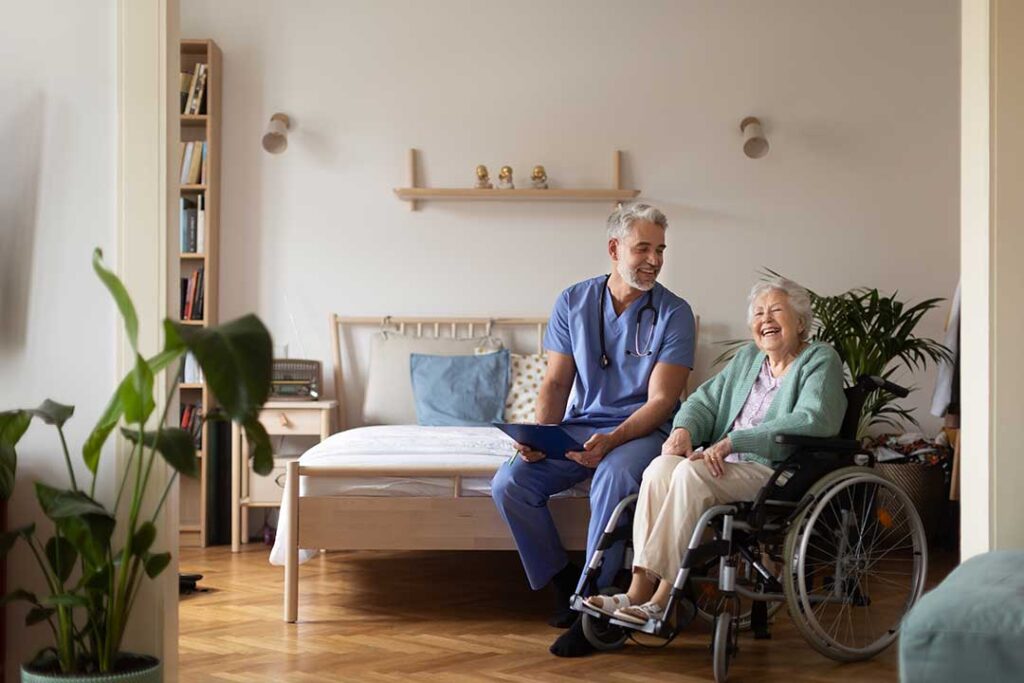An older adult smiling and chatting with a caregiver in a cozy, well-lit communal living space.