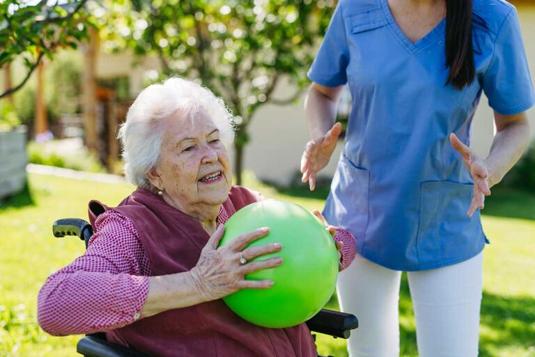 Caregiver helps an older resident walk safely through a bright assisted living hallway.